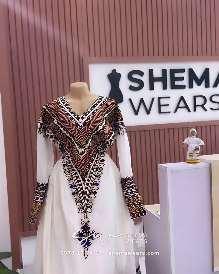 White habesha kemis on a mannequin with a deep V of brown and black tilf embroidery, long sleeves and a patterned hem