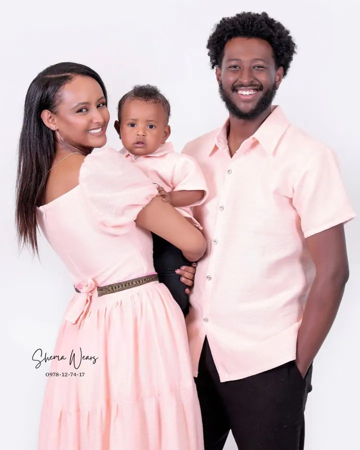 A couple holding their baby in matching blush pink — her puff-sleeve maxi dress, his short-sleeve shirt