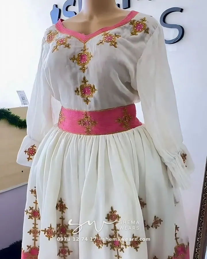 White dress with sheer long sleeves, a pink waistband and pink and gold embroidered crosses on the bodice and skirt