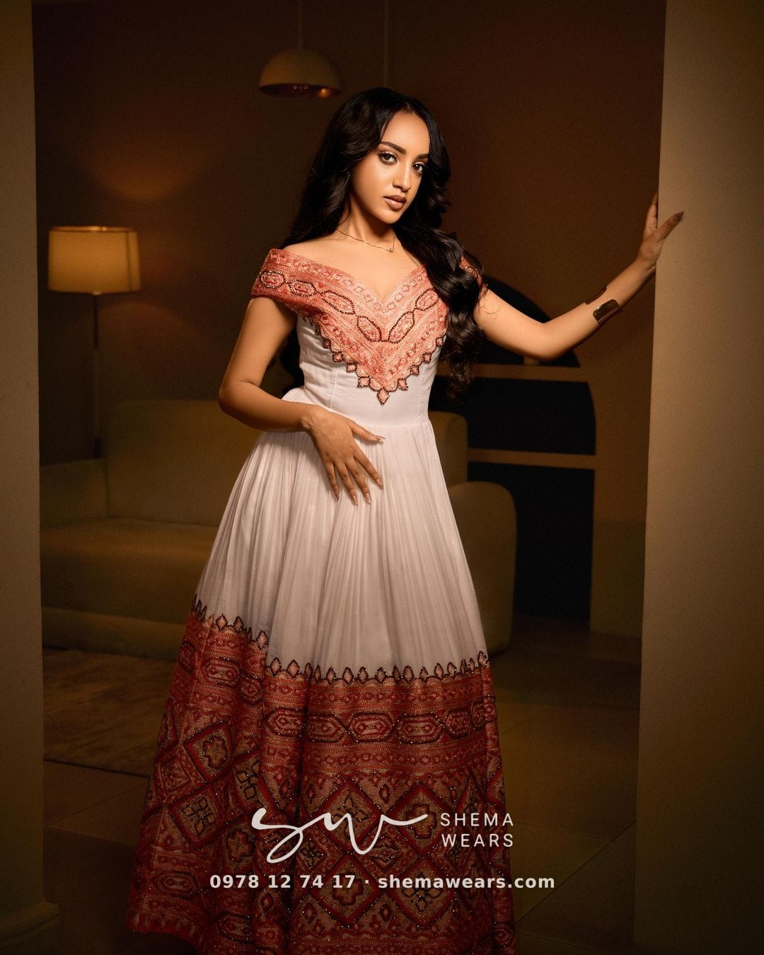 Model in a white off-shoulder habesha kemis with coral tibeb across the bodice and a wide coral tibeb skirt