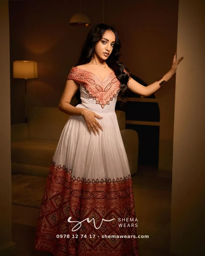 Model in a white off-shoulder habesha kemis with coral tibeb across the bodice and a wide coral tibeb skirt