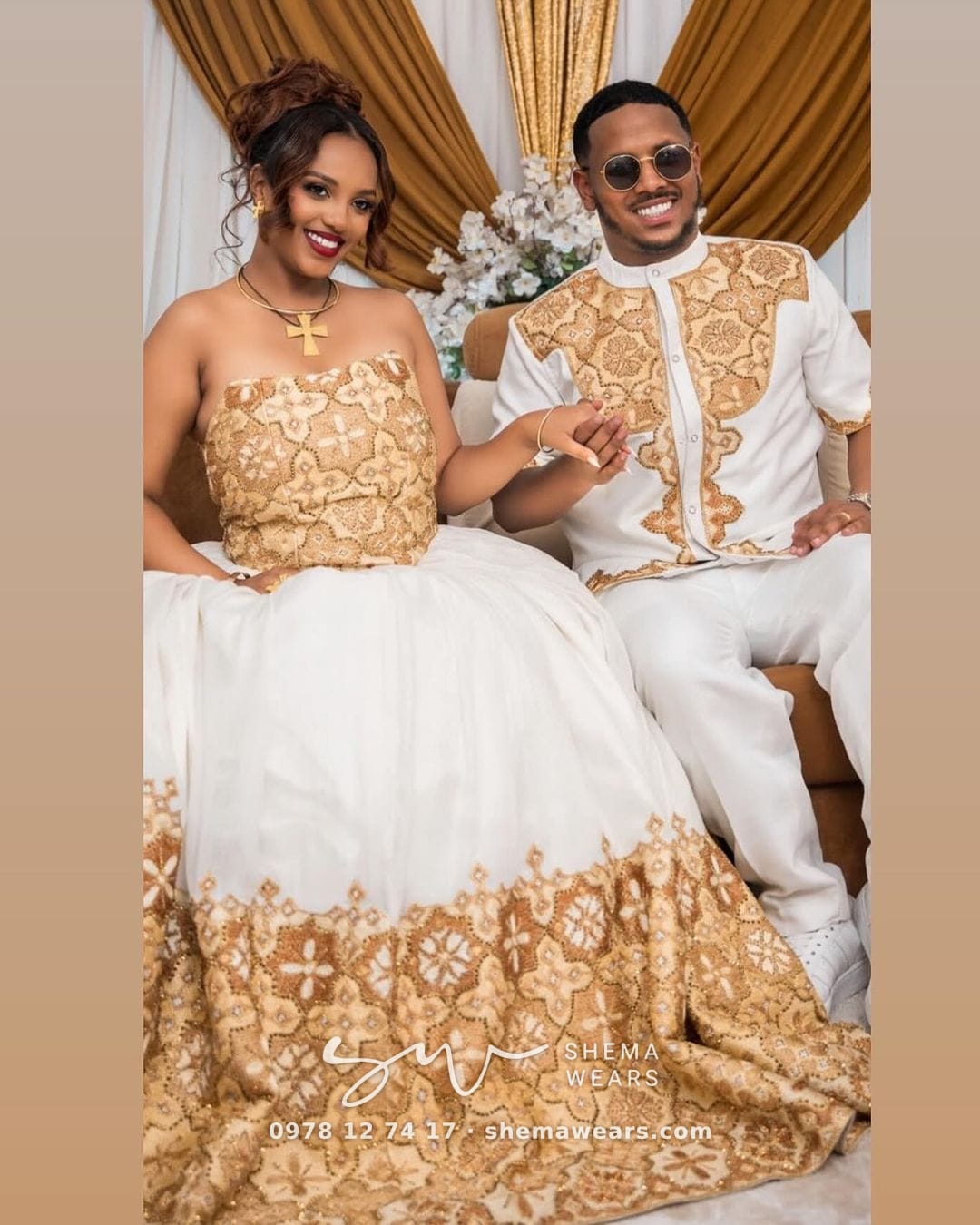 A bride in a white ballgown with a gold tibeb strapless bodice and hem, beside a groom in white with gold tibeb