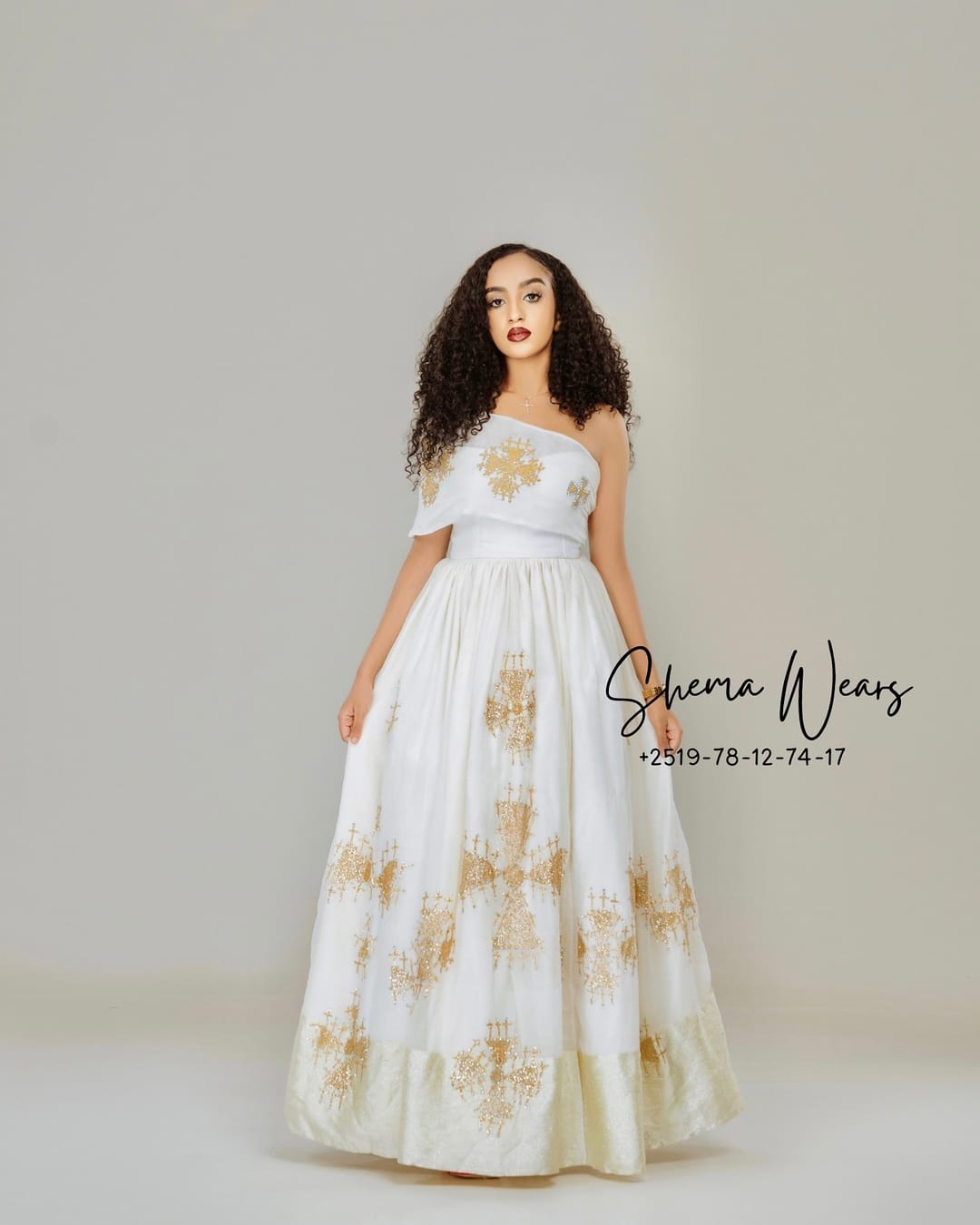 Model in a white one-shoulder habesha kemis with scattered gold embroidered crosses and a gold hem