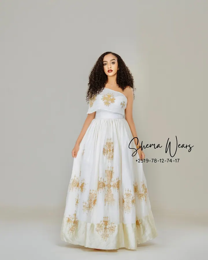 Model in a white one-shoulder habesha kemis with scattered gold embroidered crosses and a gold hem
