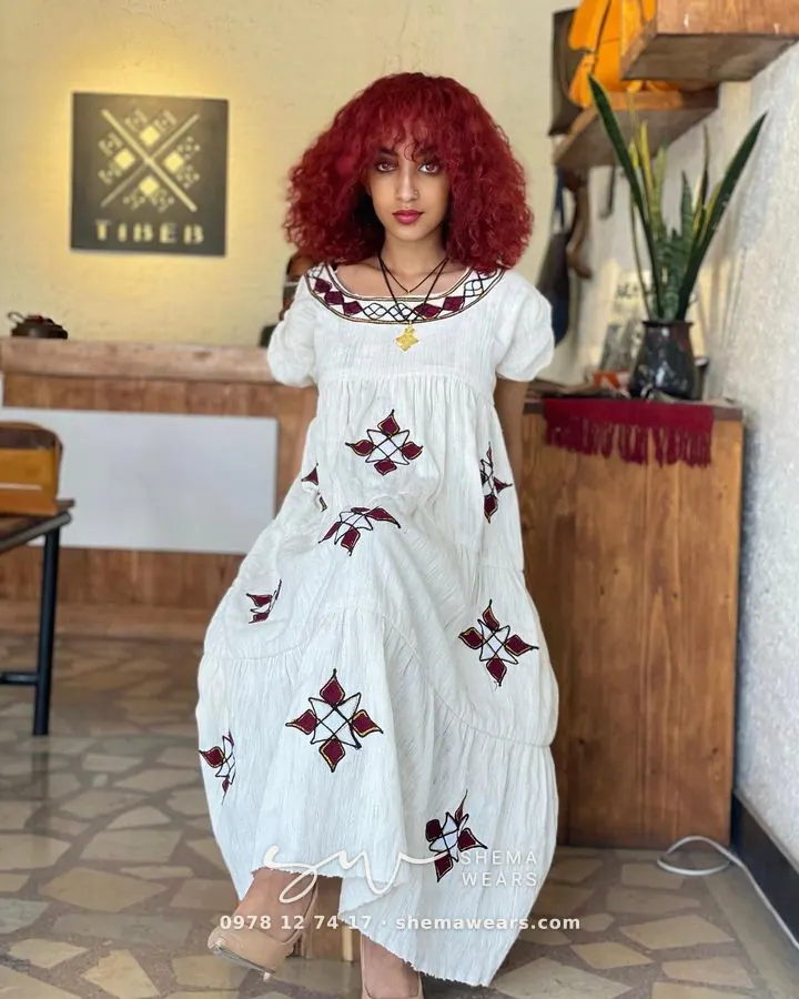 Model in a loose white habesha dress with maroon embroidery at the square neckline and scattered maroon crosses