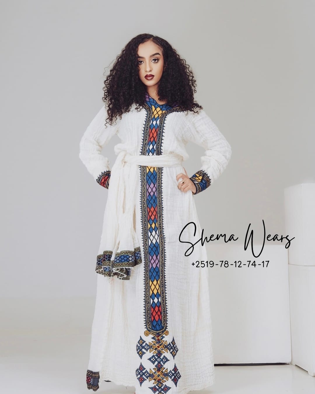Model in a white long-sleeve habesha kemis with a blue, yellow, red and black tibeb panel, cuffs and belt