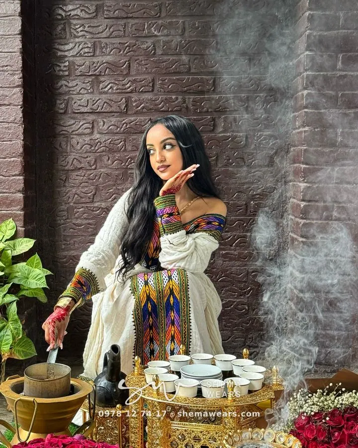 Woman at a coffee ceremony in a white off-shoulder habesha kemis with multicolour tibeb on the bodice and sleeves