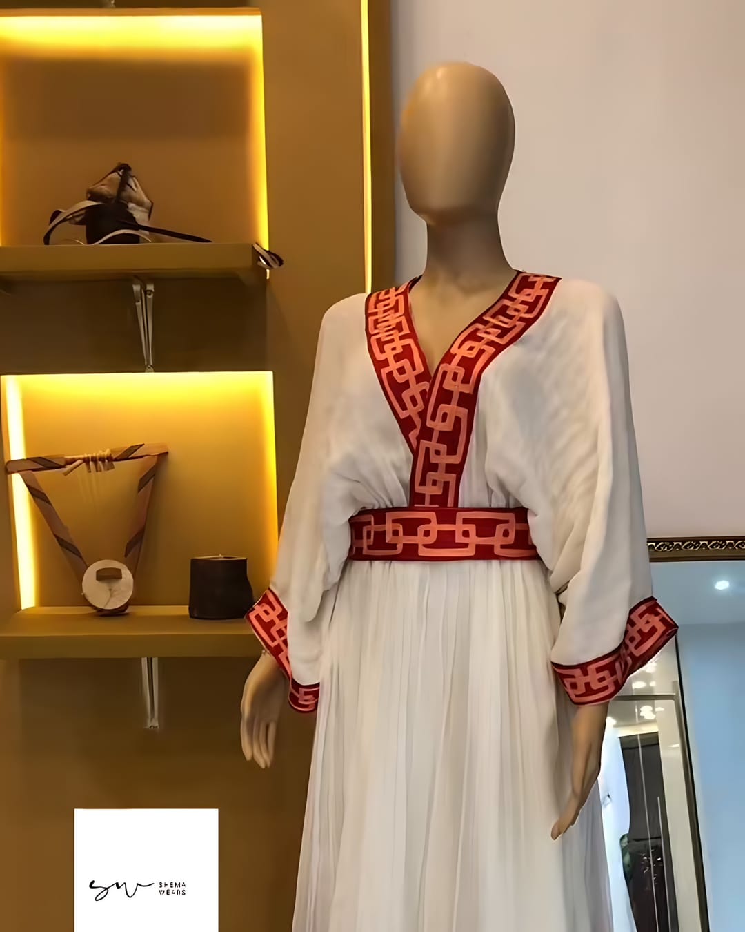 ኣብ ማኒኪን ዘሎ ጻዕዳ ሓበሻ ቀሚሽ፣ ሰፊሕ ኢድ፣ ቀይሕ ዝተኣሳሰረ ቅዲ ጥበብ ዘለዎ V ክሳድ፣ ቅናትን ኣብ ጫፉ መስመራትን ዘለዎ