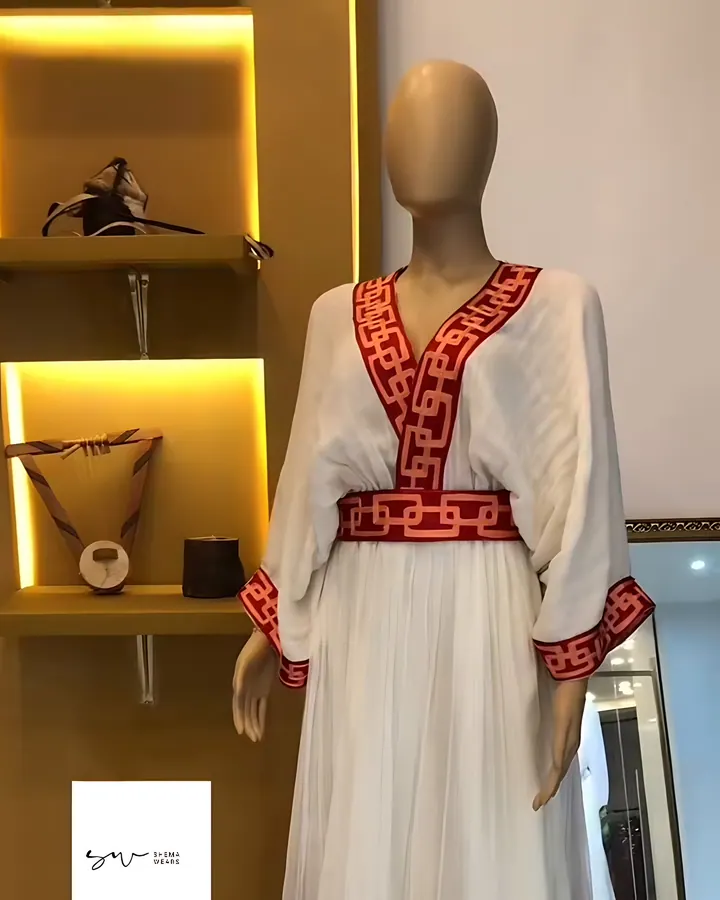 White habesha kemis on a mannequin with wide sleeves, a red key-pattern tibeb V neckline, belt and hem bands