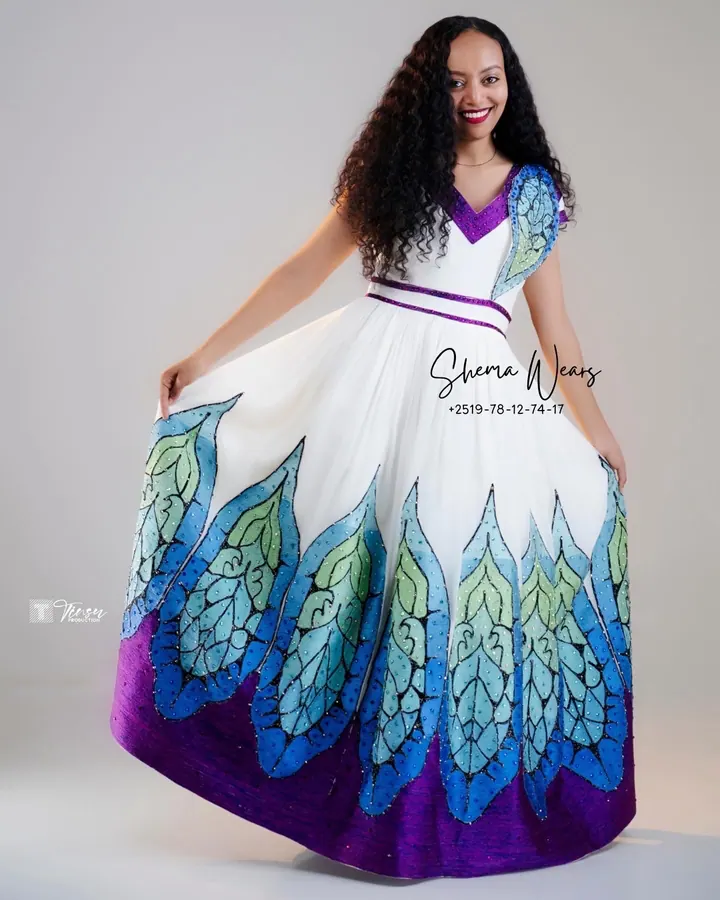 Model holding out the skirt of a white habesha kemis with large blue, green and purple peacock-feather tibeb